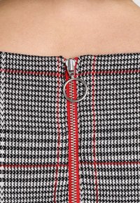 Tissu à carreaux noir et blanc avec des accents rouges ; présente une fermeture éclair en métal visible et un petit tirette circulaire à l'arrière.