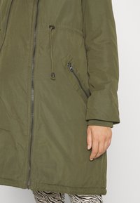 Chaqueta parka verde oliva con cremallera frontal, cordón ajustable en la cintura y bolsillo lateral con cremallera. Tiene una tela lisa y texturizada y mangas largas.