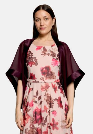 Frau mit dunklen glatten Haaren, die ein ärmelloses Kleid mit pinken und roten Blumen trägt und einen dunkelvioletten transparenten Schal über den Schultern hat.