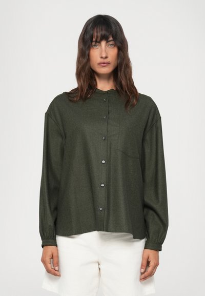 Chemise boutonnée verte foncée en tissu texturé, avec un col, des manches longues et une poche poitrine. Stylée avec un short blanc.