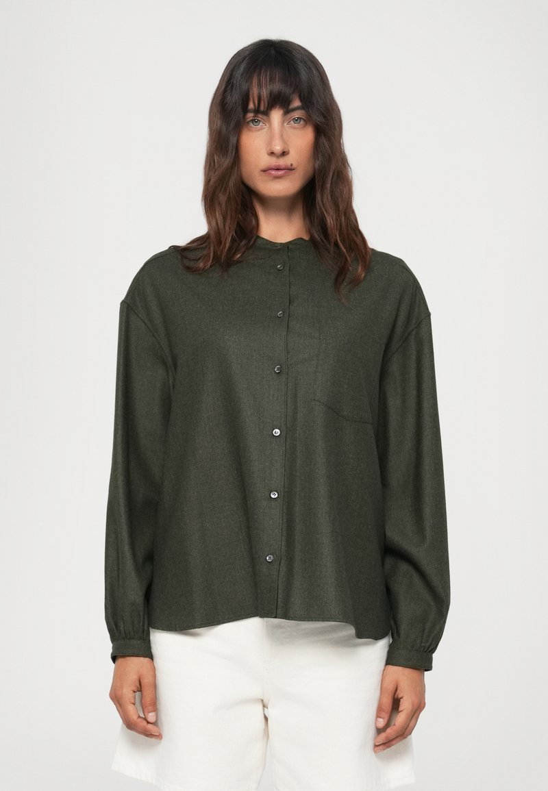 Cămașă cu nasturi de culoare verde închis, fabricată dintr-un material texturat, având guler, mâneci lungi și un buzunar pe piept. Stilizată cu pantaloni scurți albi.