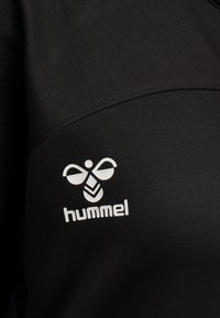 Sort sportsbeklædning med et hvidt Hummel-logo, der har et design med en bi og en pil. Materialet er glat med en let glans og subtil tekstur.
