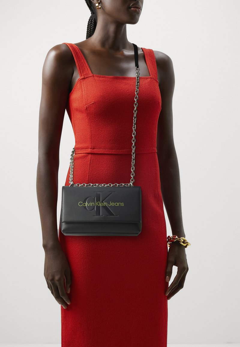 Bolso bandolera de cuero negro con logotipo embosado, cadena como asa y letras verdes, combinado con un vestido ajustado de textura roja.