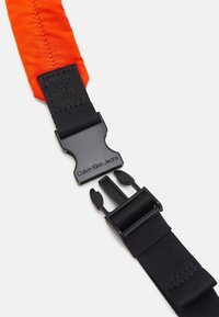 Czarny pasek nylonowy z jasnopomarańczową sekcją, z czarną plastikową klamrą z napisem "Calvin Klein Jeans." Tkanina teksturowana i gładkie okucia.