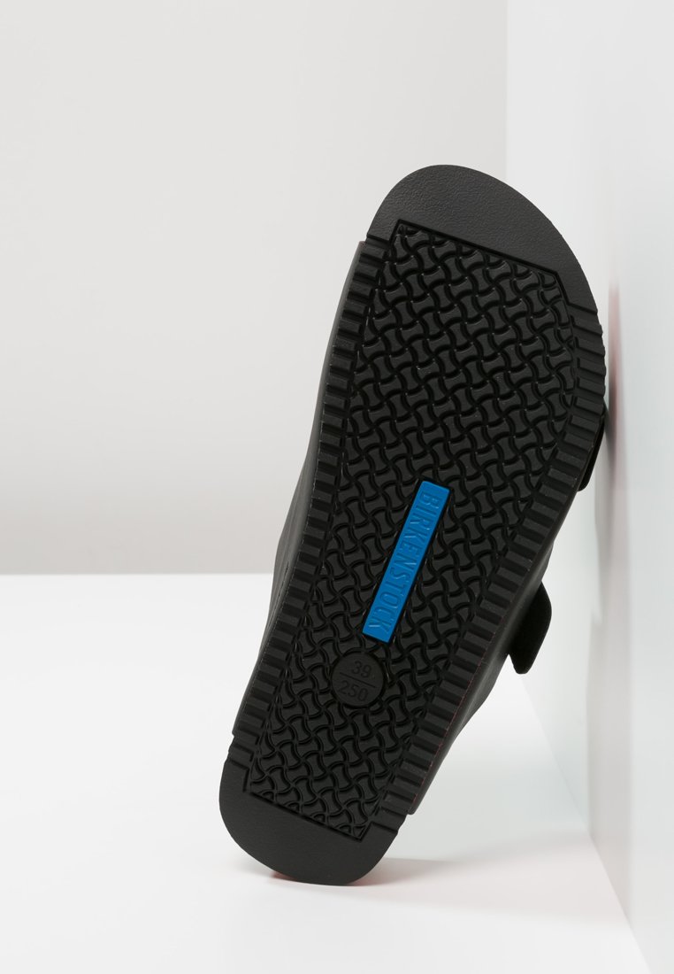 Black footwear with a textured rubber sole, featuring a blue label that reads "BIRKENSTOCK" and size markings, viewed from beneath.