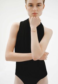 Top negro sin mangas con un cuello alto y un diseño ajustado. Modelo llevando una pulsera dorada con un pequeño charm circular en la muñeca.