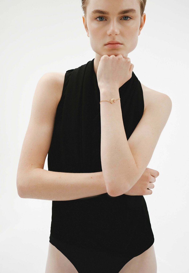 Top negro sin mangas con un cuello alto y un diseño ajustado. Modelo llevando una pulsera dorada con un pequeño charm circular en la muñeca.
