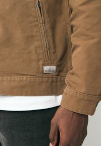 Giacca in tela marrone con tasca con cerniera, maniche con polsini e un'etichetta all'orlo. Sotto, si vede una camicia bianca.