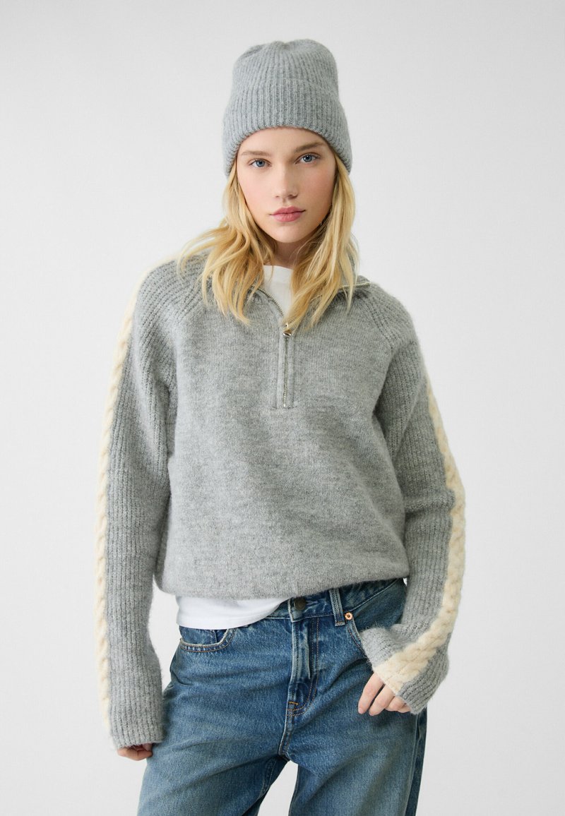 Maglione grigio con zip, lavorato a maglia con un motivo testurizzato e dettagli a treccia color crema sulle maniche, abbinato a un berretto a coste grigio coordinato e jeans blu.