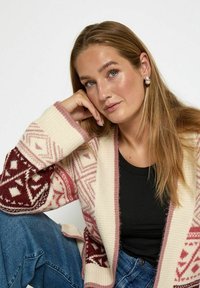 Gebreide cardigan in crème, bordeaux en roze met geometrische patronen en ribbels aan de mouwen. Gecombineerd met een zwarte top en spijkerbroek.