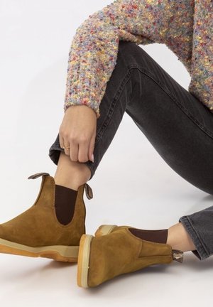 Botines de ante marrón con paneles elásticos en los costados, suelas de goma ligeras y tiradores, combinados con jeans oscuros y un suéter multicolor.