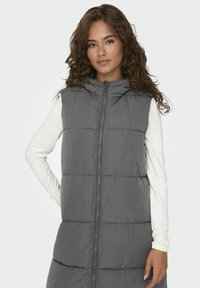 Gilet en duvet gris avec capuche, doté de coutures horizontales, fermeture éclair à l'avant et design long. Assorti à une chemise blanche à manches longues.