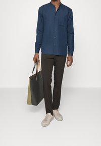 Camisa azul marino abotonada con cuello mandarín, combinada con pantalones oscuros y zapatillas claras. Sosteniendo un bolso tote bicolor en verde y negro.