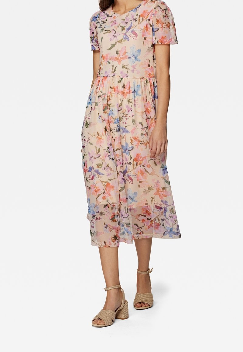 Femme portant une robe midi à manches courtes fleurie avec des fleurs rose pastel, bleu et orange, associée à des sandales beiges tissées à talons carrés.