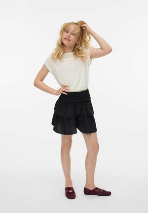 Blond meisje staat met een hand op de heup en aan haar haar, gekleed in een wit T-shirt, een zwart gelaagd rokje en bordeauxrode flats op een witte achtergrond.