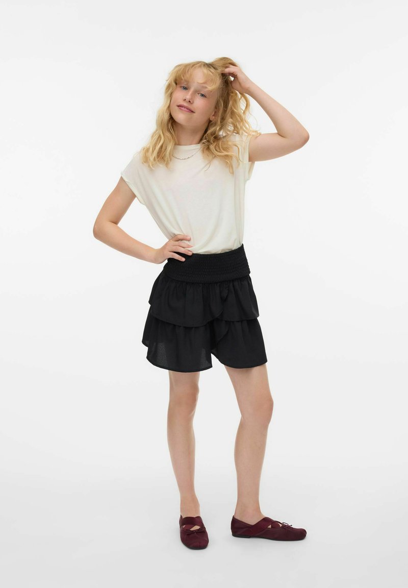 Blondine står med hånden på hoften og rører ved sit hår, iført en hvid t-shirt, sort lagdelt nederdel og burgunder flade sko på hvid baggrund.