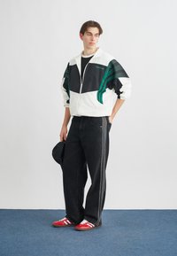 Balta un melna kabatas jaka ar zaļām svītrām, kombinēta ar plašām melnām biksēm ar sānu svītrām un sarkanām kedām. Modelis pozē pret neitrālu fonu.