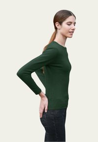 Haut vert à manches longues avec un design ajusté et un col rond, en tissu doux. Associé avec un jean noir en vue de côté.