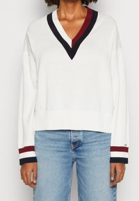 Weißer V-Ausschnitt-Pullover mit Marineblau- und Weinrot-Streifenakzenten am Ausschnitt und den Bündchen, mit cropped Design und geripptem Saum. Kombiniert mit hellblauen Jeans.