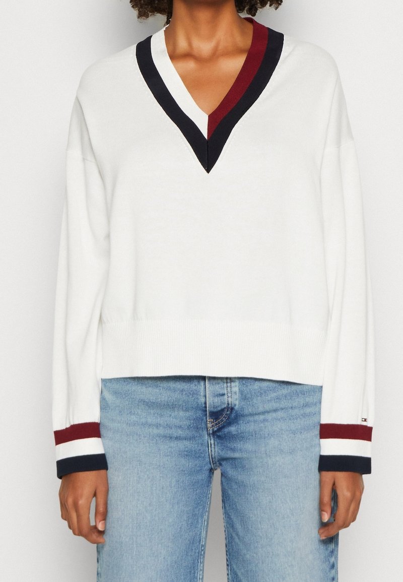 Weißer V-Ausschnitt-Pullover mit Marineblau- und Weinrot-Streifenakzenten am Ausschnitt und den Bündchen, mit cropped Design und geripptem Saum. Kombiniert mit hellblauen Jeans.