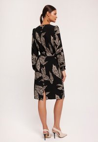 Czarna sukienka z liściastym wzorem w kolorze kremowym, długie rękawy i pasek w talii; rozcięcie z tyłu. Połączona z beżowymi szpilkami.