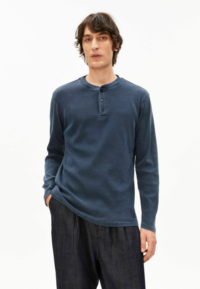 Marineblaues Langarm-Henley-Shirt mit strukturierter Strickoptik, Drei-Knopf-Leiste und lockerem Schnitt. Kombiniert mit dunklen Jeanshosen.