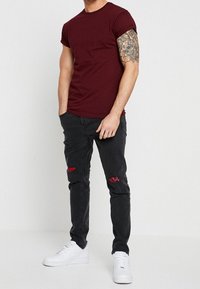 Camiseta de algodón burdeos combinada con jeans negros con detalles desgastados y parches de cuadro escocés rojo; zapatillas blancas completan el conjunto.