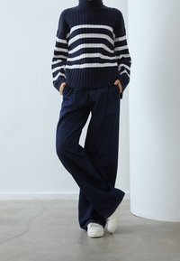 Pull rayé bleu marine à texture côtelée et col haut, associé à un jean denim foncé à jambes larges et des baskets blanches.