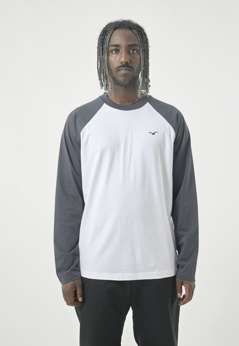 Langärmliges Raglan-Shirt mit weißem Oberteil und dunkelgrauen Ärmeln. Aus Baumwollmaterial, Rundhalsausschnitt, kleines Logo auf der Brust und lässige Passform.