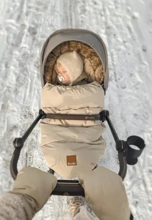 Nukkuva vauva beigessä rattaita, pukeutuneena lämpimästi ja hupullisesti, työnnetään lumisella polulla henkilö toimesta, joka käyttää beigen värisiä käsineitä.