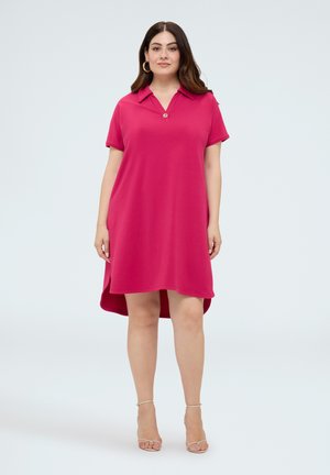 Kvinde iført en kortærmet pink polo-kjole med høj-lav kant, beige hælede sandaler og gyldne hoop-øreringe, stående mod en ensfarvet baggrund.