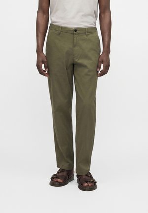 Personne portant un pantalon droit vert olive, une chemise blanche et des sandales marron à bouts ouverts, debout devant un fond blanc uni.