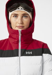 Chaqueta acolchada roja, negra y blanca con cremallera, cuello alto y logo en el pecho. Presenta una capucha ajustada y una textura suave y brillante.