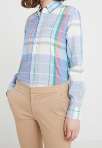Camicia a maniche lunghe a cuadros blu chiaro con righe verde, rosa e bianco; colletto button-down; indossata con pantaloni attillati beige.
