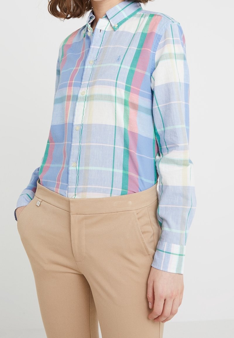 Camicia a maniche lunghe a cuadros blu chiaro con righe verde, rosa e bianco; colletto button-down; indossata con pantaloni attillati beige.