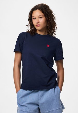 Femme aux cheveux bouclés portant un T-shirt bleu marine avec un petit cœur rouge sur la poitrine gauche et un short en jean bleu clair.
