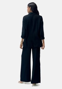 Zwart textuuroutfit bestaande uit een langemouwen shirt en wijde broek. Het shirt heeft een relaxte pasvorm en beide stukken zijn gemaakt van een gekreukt materiaal.