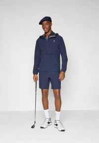 Marineblå atletisk genser med halv glidelås og hette, kombinert med matchende shorts. Hvite joggesko og golfkølle fullfører sportsantrekket.