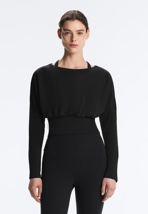 Žena s tmavými vlasmi zviazanými dozadu má na sebe voľný čierny crop top s dlhým rukávom cez priliehavú čiernu športovú podprsenku a legíny s vysokým pásom.