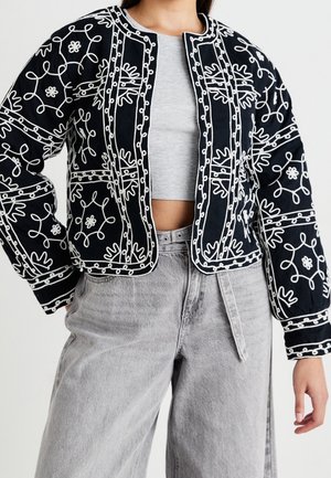 Czarna kurtka z długim rękawem z białymi haftowanymi kwiatowymi i abstrakcyjnymi wzorami, noszona na szarym, krótko skrojonym prążkowanym topie i jasnoszarych spodniach z wysokim stanem.