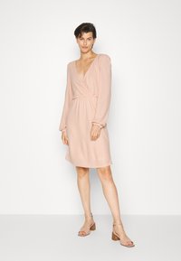 Vestido envolvente rosa com mangas longas, decote em V, tecido texturizado, cintura franzida e comprimento acima do joelho. Combinado com tacones de tiras claras.