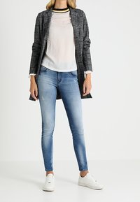 Langer, grauer Karomantel über einer transparenten weißen Bluse, kombiniert mit hellblauen Skinny Jeans und weißen Sneakers. Hat einen schwarz-gelben Kragen.