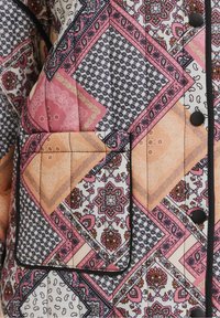 Quiltet jakke i pastelfarver med patchwork-design, der har indviklede mønstre. Sort piping afgrænser lommen og knapperne.