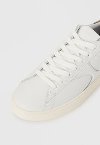 Sneaker in pelle bianca con accenti texture, dotato di lacci bianchi piatti, punta arrotondata e suola in gomma color crema. Nome del marchio visibile.