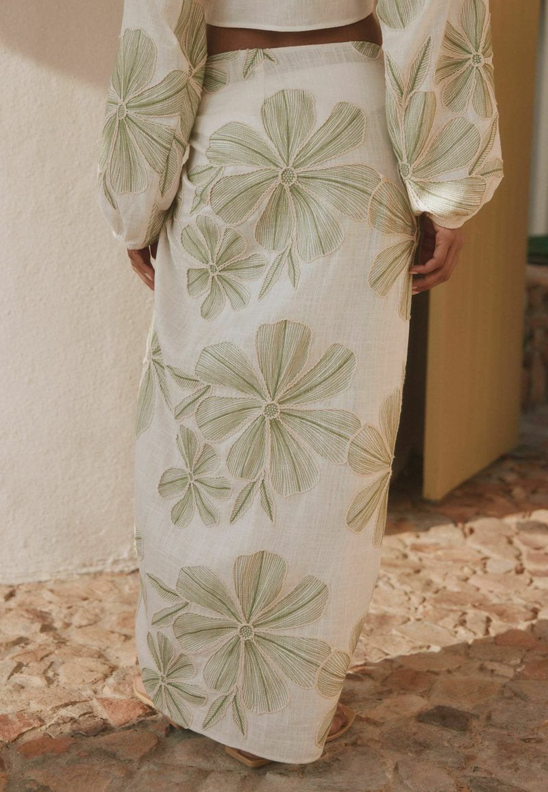 Kobieta ubrana w białą spódnicę i bluzkę z dużym zielonym haftem kwiatowym, stojąca na kamiennej podłodze obok beżowej ściany i otwartych drzwi.