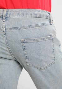 Jeans de mezclilla de un azul claro, que presentan un bolsillo trasero con detalles de costura, textura de tela visible y una cintura de altura media.