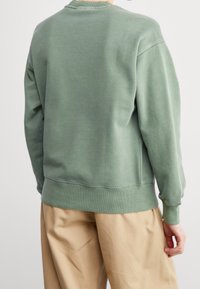 Felpa verde in cotone con polsini e orlo a coste, caratterizzata da un design semplice e una consistenza morbida, indossata con pantaloni beige.