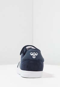 Navy suede sneaker with white rubber sole, featuring three white stripes and a logo on the heel. Soft texture, low-profile design.