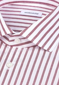 Chemise blanche à rayures rouges, dotée d’un col boutonné et d’un seul bouton blanc sur la patte de boutonnage avant. Étiquette visible à l'intérieur du col.