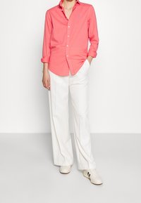 Korallfärgad skjorta med knappar och långa ärmar samt en liten blå logotyp, parat med vita vida byxor och vita sneakers.